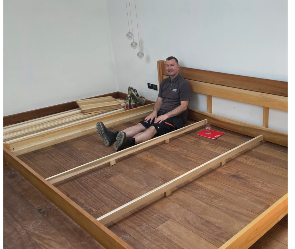 Man sitting on an unfinished wooden bed frame.