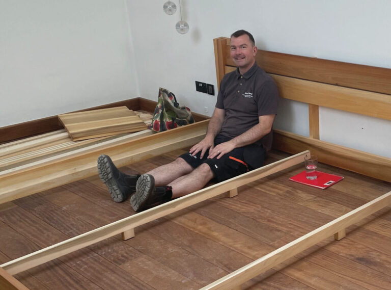 Man assembling wooden bed frame, smiling indoors.