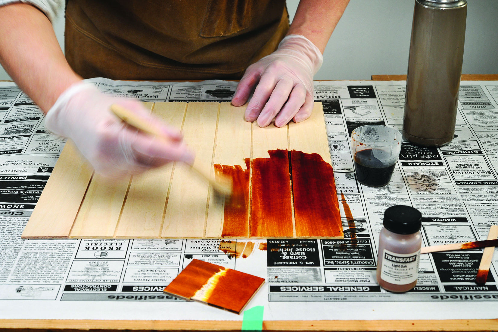 Person staining wood with brown dye on newspaper.