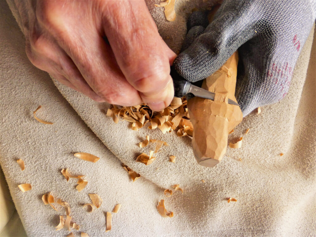 Hands carving wood with chisel and gloves.