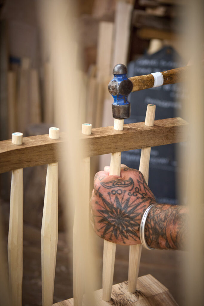 Tattooed craftsman assembling wooden chair with hammer.