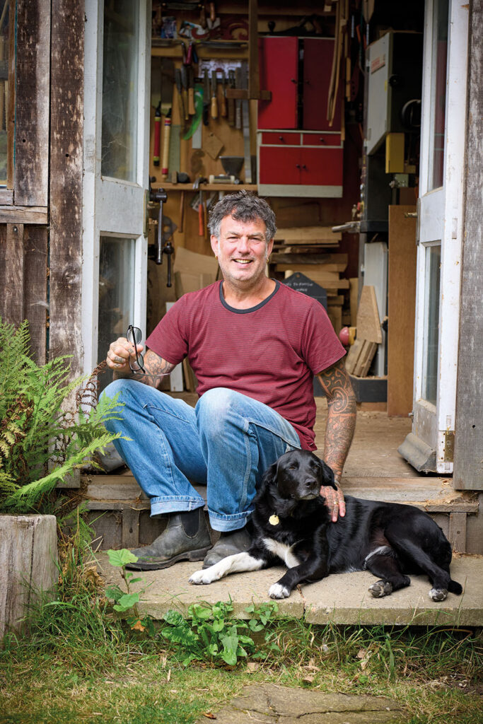 Jason Mosseri sitting with dog outside rustic workshop.