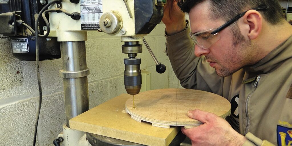 Man operating pillar drill on wooden board.