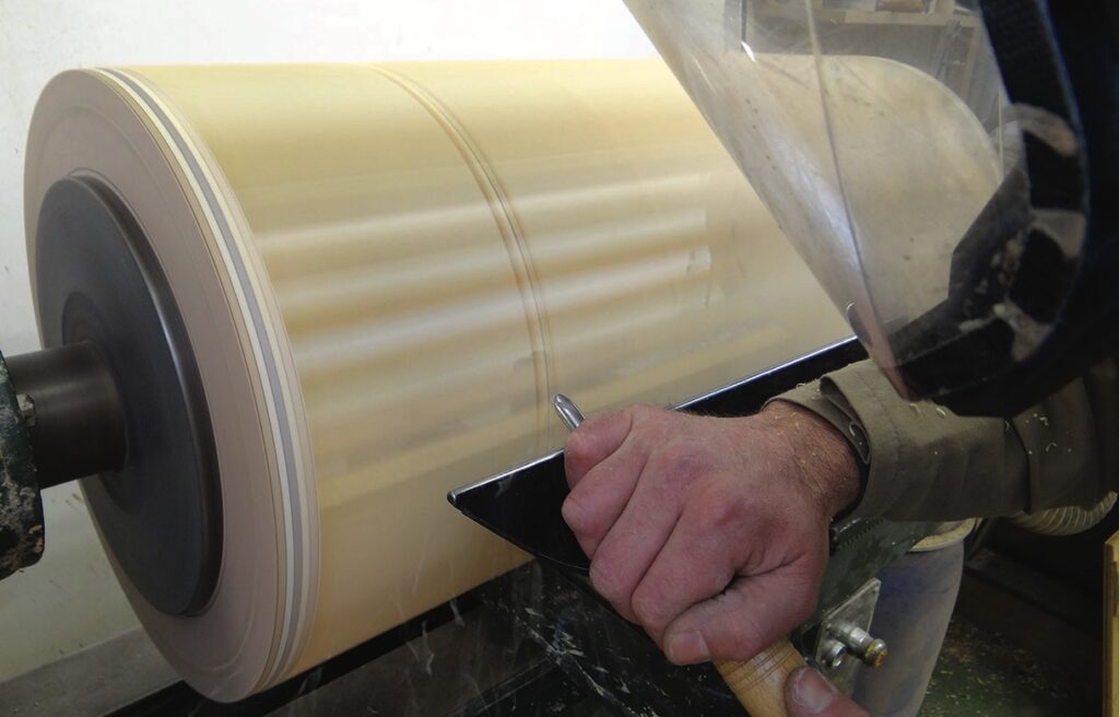 Woodturner shaping a large spinning cylinder on lathe.