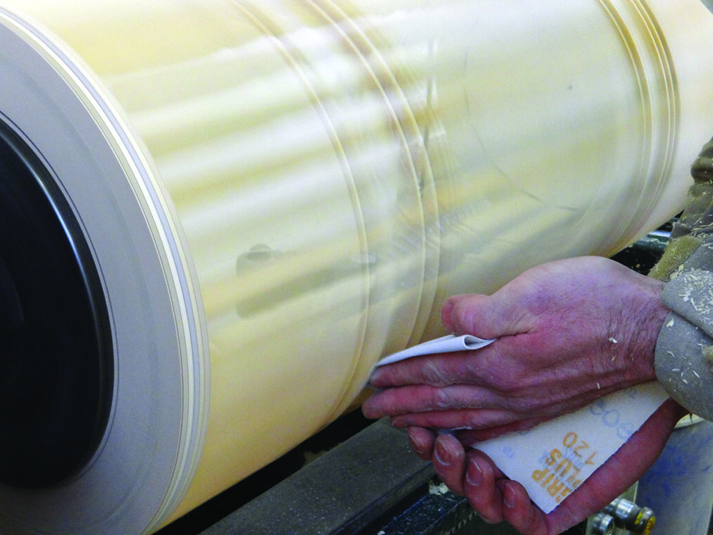 Person sanding large wooden cylinder on lathe.