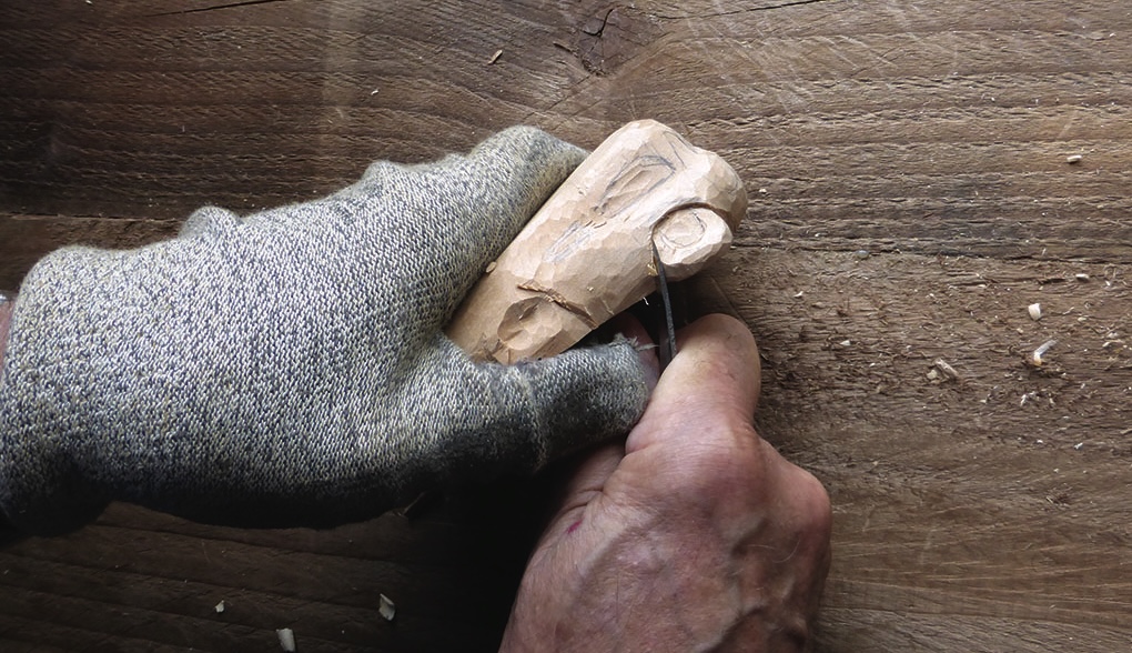 Gloved hand carving wood with a small knife.
