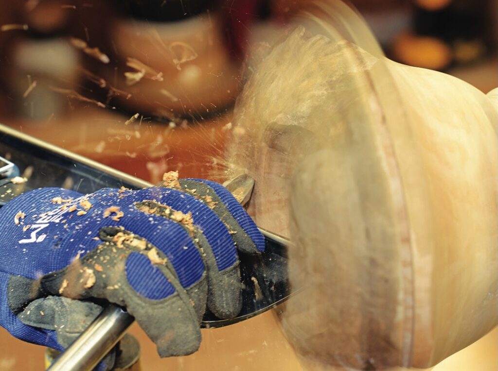 Gloved hand woodturning on lathe with flying shavings.