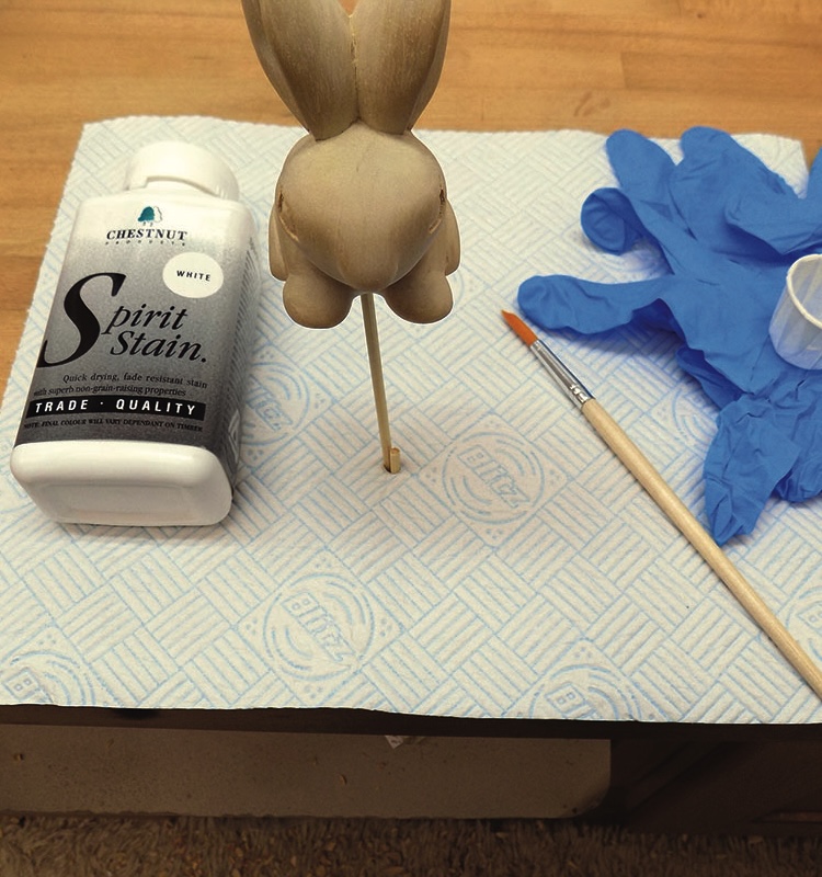 Wooden bunny with stain, brush, gloves on table.