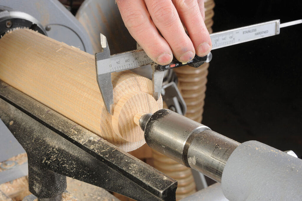 Measuring turned wood piece with caliper on lathe.