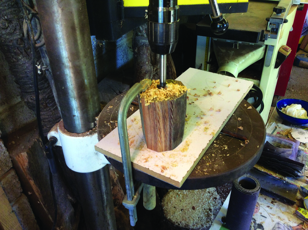 Drill press cutting into wooden block in workshop.