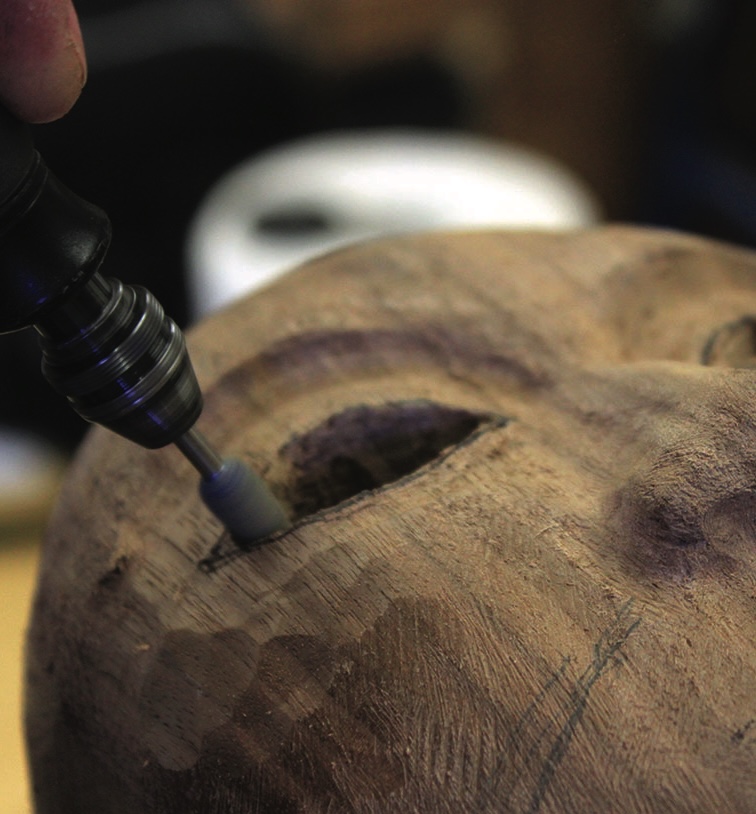 Hand using rotary tool on wooden sculpture face.